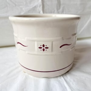 Puede incluir: Porta utensilios de cerámica color crema con un diseño decorativo. El soporte presenta una banda de formas rectangulares con un motivo floral y líneas curvas en color burdeos intenso. Una línea burdeos rodea la base.