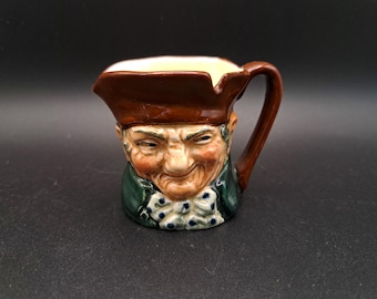 May include: A small, ceramic pitcher shaped like a smiling man's head. The pitcher has a brown hat, a green jacket, and a white bow tie with blue dots. The handle and the inside of the pitcher are brown.