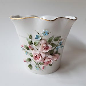 Puede incluir: Un florero de porcelana blanca con un borde acampanado y festoneado con un acabado dorado. El exterior está decorado con rosas rosadas de cerámica en relieve, hojas verdes y pequeñas flores azules aplicadas sobre la superficie frontal del recipiente.