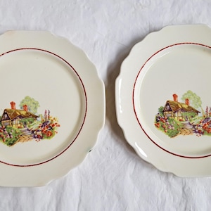 May include: Two white ceramic plates with scalloped edges and a red rim. Each plate features a cottage with a thatched roof and a garden scene in the centre.