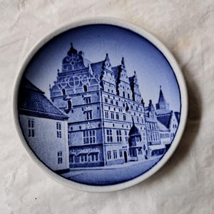 Può includere: Un piatto in ceramica blu e bianco con un'illustrazione dettagliata di un edificio storico in una città europea. L'edificio ha un tetto a punta e molte finestre.