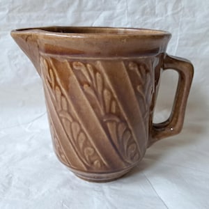 May include: A brown ceramic pitcher with a handle and a spout. The pitcher has a textured, embossed design of leaves and diagonal lines. The pitcher is likely vintage and suitable for serving beverages.