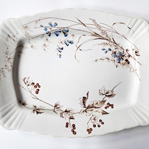 May include: A white rectangular serving platter with a scalloped edge. The platter is decorated with brown and blue floral and botanical designs. The design includes sprigs, leaves, and berries. The platter is likely made of ceramic or porcelain.