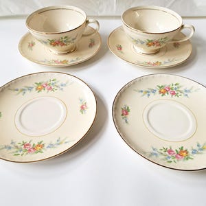 May include: A set of vintage cream-colored teacups and saucers with floral designs. The set includes two teacups, two saucers, and two oval-shaped serving plates. Each piece features a gold trim and a delicate pattern of pink, yellow, and blue flowers.
