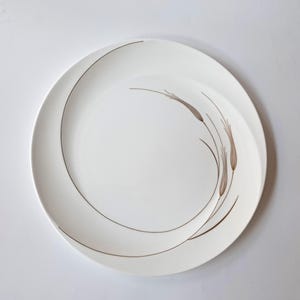 May include: A round white ceramic dinner plate featuring a minimalist brown line drawing of wheat stalks arranged in a sweeping, curved pattern that follows the circular edge of the plate, leaving the centre of the dish clean and white.