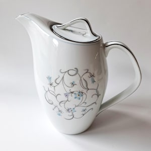 May include: A white porcelain coffee pot featuring a delicate gray swirling vine pattern accented with small light blue flowers. The pot includes a matching lid with a handle, a curved spout, and a silver-toned trim along the rim and handle.
