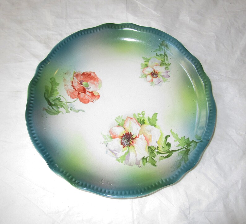 May include: A white ceramic plate with a scalloped edge and a green and blue glaze. The plate is decorated with hand-painted flowers in shades of pink, white, and green.