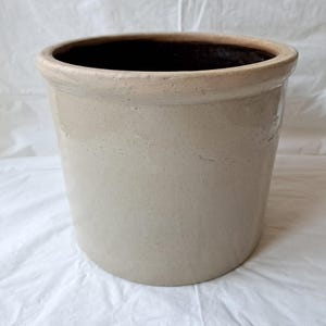 May include: A cylindrical stoneware crock featuring a smooth, light beige exterior finish and a thick, rounded rim. The interior of the vessel is dark brown, contrasting with the lighter outer surface. The pot has a simple, straight-sided design.
