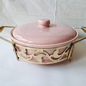 May include: A round, speckled pink ceramic casserole dish with a matching lid, set inside a decorative gold-tone metal cradle. The cradle features a cut-out leaf pattern and two gold-tone wire coil handles on opposite sides of the dish.