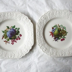 Könnte beinhalten: Zwei weiße Keramikteller mit aufwendigen Reliefmustern an den Rändern. Ein Teller zeigt ein gemaltes Design aus Pflaumen, Trauben und Himbeeren. Der andere Teller zeigt ein gemaltes Design aus einer Birne, Trauben und Himbeeren.