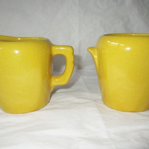 May include: A set of two yellow ceramic creamer and sugar containers. The creamer has a handle and a spout. The sugar container has two small handles.
