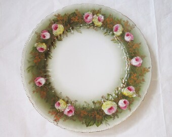 May include: A white ceramic plate with a scalloped edge, decorated with a wreath of pink and yellow roses, green leaves, and gold accents. The plate has a glossy finish and a gold rim.