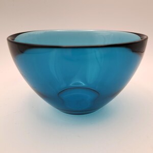 May include: A teal-colored glass bowl with a rounded shape. The rim of the bowl is outlined with a dark, almost black, band. The bowl has a smooth, glossy finish and a simple, elegant design. The bowl is empty.
