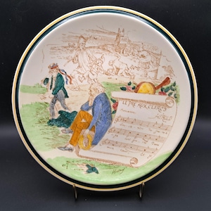 Könnte beinhalten: Dekorative Keramikplatte mit detaillierter Illustration. Die Platte zeigt eine Szene mit Figuren, Noten und Text. Der Rand hat einen grünen und goldenen Rand. Die Platte wird auf einem Metallständer ausgestellt.