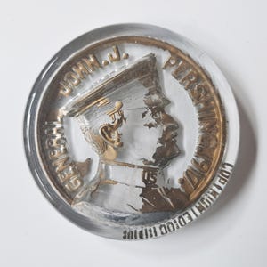 May include: A clear, circular commemorative coin featuring a profile of a man in a military uniform. The coin has gold and silver tones, with the text "JOHN J. PERSHING 1917" and "GENERAL" around the edge. The coin is set in a clear, glass-like material.