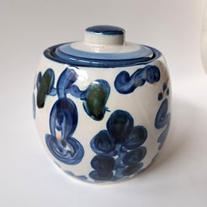 May include: A ceramic jar with a lid, featuring a white base adorned with blue floral patterns and accents. The lid has concentric blue stripes. The jar's design includes dark blue and green brushstrokes, creating a unique, handcrafted appearance.