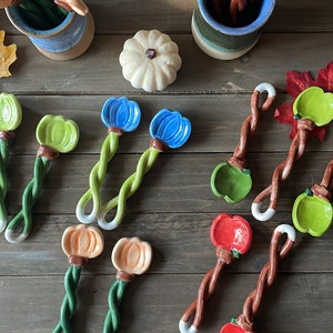 May include: A collection of ceramic spoons with colorful fruit and flower designs. The spoons feature twisted green stems and white tips. The fruit and flower colors include orange, blue, green, and red. The spoons are arranged on a wooden surface.