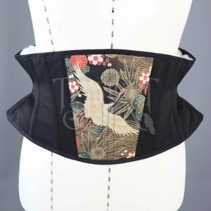 Puede incluir: Corsé negro con un panel decorativo que presenta una grulla, flores y follaje en tonos dorados, rojos y verdes. El corsé se exhibe en un maniquí, mostrando su forma y diseño.