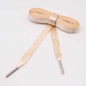 May include: A pair of beige satin shoelaces with silver metal aglets. The laces are tied in a bow.