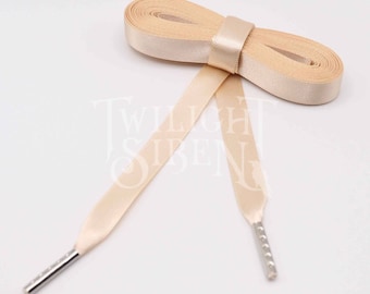 Puede incluir: Un par de cordones de zapatos de satén beige con aglets de metal plateado. Los cordones están atados en un lazo.