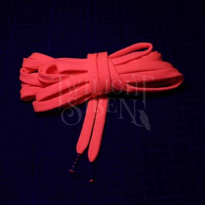 May include: Vibrant coral shoelaces, knotted and bundled, set against a dark blue backdrop. The laces feature a flat, woven texture and are finished with orange and blue aglets. A fashionable accessory for trainers or shoes.