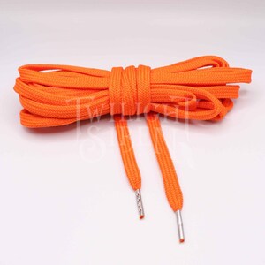 May include: A pair of bright orange shoelaces with silver aglets. The shoelaces are made of a woven material and are tied together in a knot.