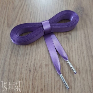 Puede incluir: Cordones de zapatos de satén morado con aglets plateados. Los cordones están atados en un lazo y están sobre una superficie de madera.