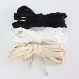 May include: Three pairs of shoelaces in black, white, and beige. The laces are made of a flat, woven material and have metal aglets at the ends.