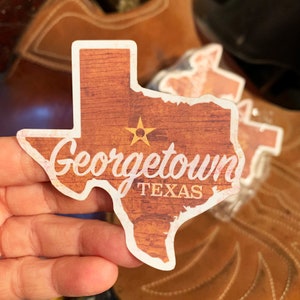 May include: A brown wooden sticker shaped like the state of Texas with the text "Georgetown, TEXAS" and a gold star in the center.
