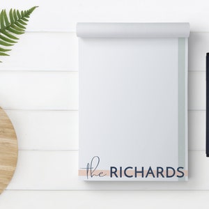 May include: A white notepad with a light pink and dark blue stripe at the bottom. The notepad has the text "the RICHARDS" printed on the bottom stripe. A black pen is leaning against the notepad.
