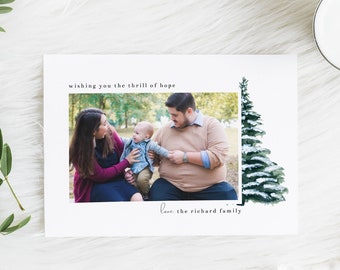 Puede incluir: Una tarjeta de Navidad con una familia de tres, una mujer, un hombre y un bebé, de pie frente a un árbol de hoja perenne nevado. La tarjeta dice "Wishing you the thrill of hope" y "Love, the Richard family".