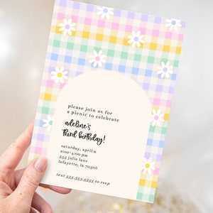 Puede incluir: Una invitación con estampado de cuadros vichy en tonos pastel rosa, azul, verde y amarillo con margaritas blancas. La invitación dice "Por favor, únase a nosotros para un picnic para celebrar el tercer cumpleaños de Adeline! Sábado, 6 de abril, 2:00-4:00 pm, 555 Julie Lane, Lafayette, LA 70508, Texto 555-555-5555 para RSVP."
