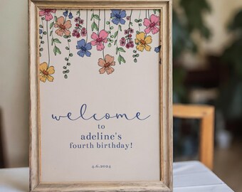 May include: A rectangular wooden frame containing a sign with a cream background, decorated with hanging colorful flowers in pink, yellow, and blue. The text reads: welcome to adeline's fourth birthday! 4.6.2024 in dark blue script and print.