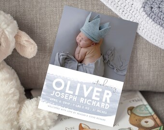 May include: A baby announcement card with a watercolor background in shades of blue. The card features a photo of a newborn baby wearing a crown and the text "introducing OLIVER JOSEPH RICHARD APRIL 6, 2019 | 7 LBS 2 OZ | 21 INCHES WELCOMED WITH LOVE BY REAGAN & STEVE RICHARD".