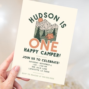 A hand is holding a birthday party invitation invitation. The invitation read "Hudson is ONE happy camper" in large text. There is an image of a camp ground with a tent, trees, and mountains in the background.