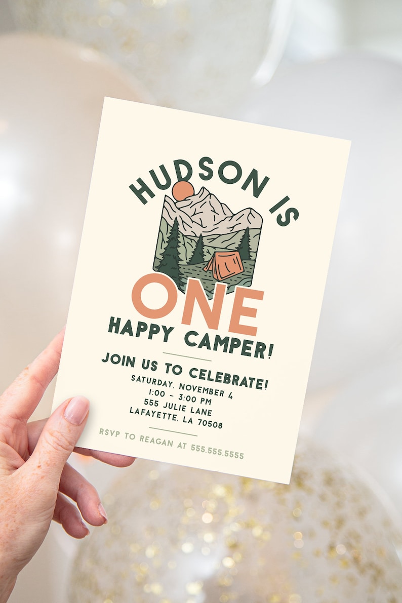 A hand is holding a birthday party invitation invitation. The invitation read "Hudson is ONE happy camper" in large text. There is an image of a camp ground with a tent, trees, and mountains in the background.