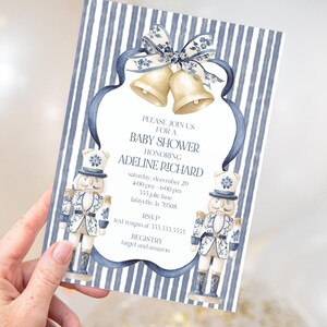 Puede incluir: Invitación a un baby shower con un fondo a rayas azules y blancas. La invitación presenta un diseño de acuarela con campanas, cintas y soldados cascanueces. El texto incluye el nombre de la homenajeada, la fecha, la hora, el lugar y la información de RSVP.
