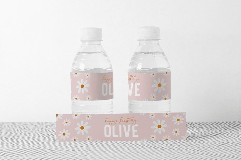 May include: Two clear plastic water bottles with pink and white daisy patterned labels that say "Happy Birthday Olive". There is also a matching label with the same pattern that says "Happy Birthday Olive".