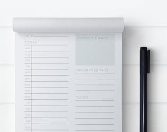 May include: A white notepad with a daily planner and to-do list. The notepad has a black pen next to it. The notepad has the words "DATE", "TODAY", "TODAY'S GOALS", "PRIORITIZE THIS", and "TO DO" printed on it.