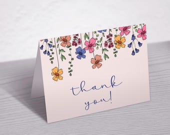 May include: A folded, light pink card with a floral design and the words "thank you!" in navy blue script. The floral design features hanging flowers in shades of yellow, pink, orange, and blue with green stems.