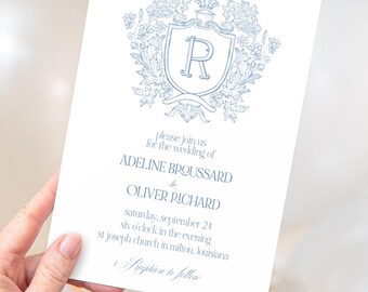 May include: A white wedding invitation with blue floral and crest design. The invitation includes the names Adeline Broussard and Oliver Richard, along with the wedding date, time, and location. The invitation is held by a hand.