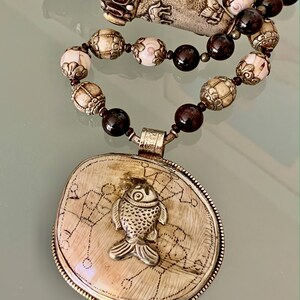 Puede incluir: Un collar de plata y hueso con un colgante que presenta un diseño de pez. El collar está hecho con cuentas de color rojo oscuro y cuentas blancas y doradas.