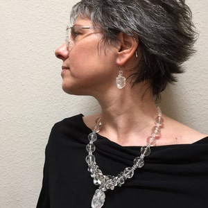 May include: A clear crystal necklace and matching earrings are displayed. The necklace features a variety of clear crystal beads and a large, irregular crystal pendant. The earrings are also made of clear crystals. The jewelry is worn against a black top.