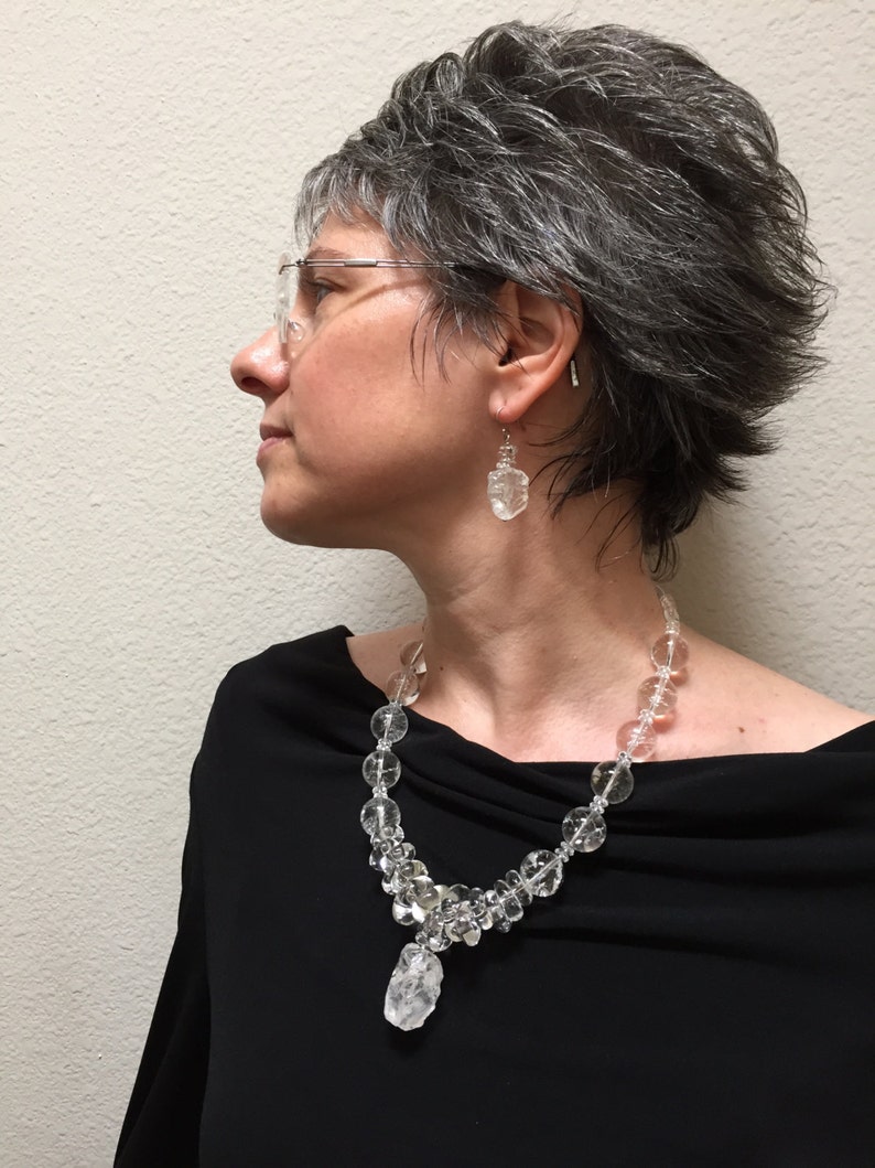 May include: A clear crystal necklace and matching earrings are displayed. The necklace features a variety of clear crystal beads and a large, irregular crystal pendant. The earrings are also made of clear crystals. The jewelry is worn against a black top.