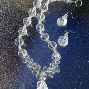 May include: A clear quartz necklace and matching earrings set. The necklace features large, round beads and a large, irregular quartz pendant. The earrings have a similar irregular quartz shape. The jewelry is displayed on a reflective, dark blue surface.