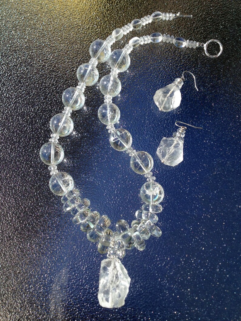 May include: A clear quartz necklace and matching earrings set. The necklace features large, round beads and a large, irregular quartz pendant. The earrings have a similar irregular quartz shape. The jewelry is displayed on a reflective, dark blue surface.