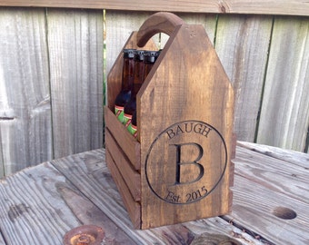 May include: Wooden six-pack beer carrier with a handle. The carrier is stained brown and has a circular cutout with the text "BAUGH B Est. 2015" engraved in it.