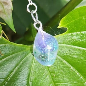 Puede incluir: Un collar de cadena de metal plateado con un pequeño colgante en forma de lágrima. El colgante está hecho de un material translúcido e iridiscente con tonos azulados, morados y verdes, que contiene finas partículas de purpurina.