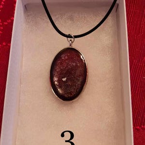 May include: An oval pendant necklace with a dark red, textured center and a silver-colored border. The pendant hangs from a black cord and is displayed in a white box. The number "3" is printed in black below the pendant.