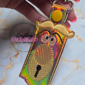 May include: A key chain with a whimsical design. The key chain features a gold embroidered mustache and a lock design with a keyhole. The key chain has a holographic background with a yellow button and a silver clasp.